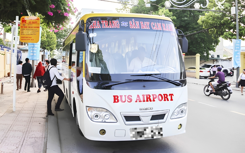 Passengers boarding a white airport shuttle bus labeled "Nha Trang - Cam Ranh Airport" on a street in Nha Trang, Vietnam.