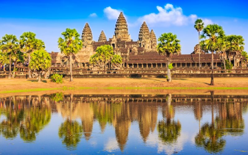 Angkor Wat temple in Cambodia with palm trees and reflection in water under a bright blue sky