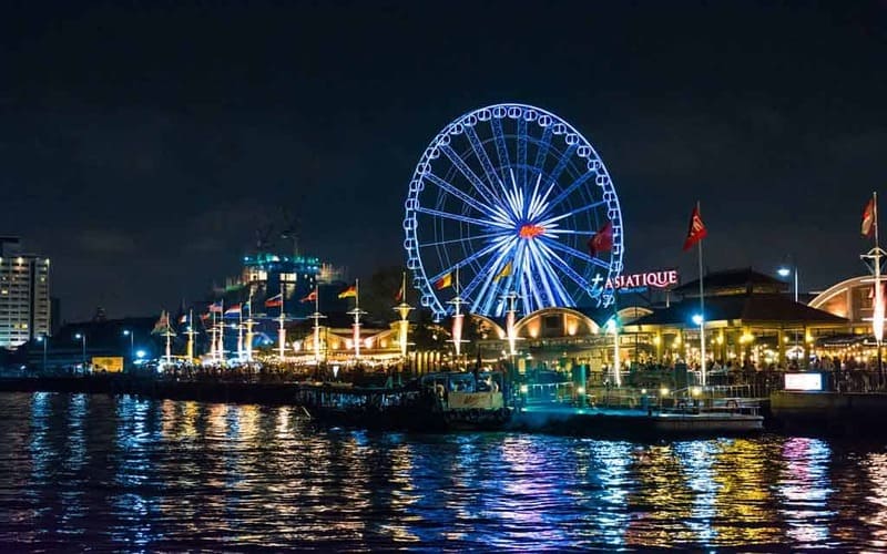 Night view of Asiatique The Riverfront in Bangkok, Thailand, featuring a brightly lit Ferris wheel reflecting on the Chao Phraya River.