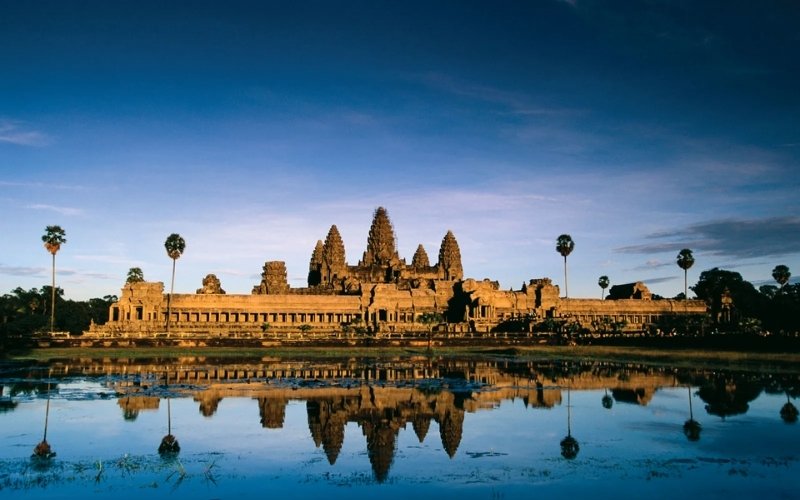 A panoramic view of Angkor Wat temple in Cambodia, reflected beautifully in a still water pond under a clear blue sky at sunrise.
