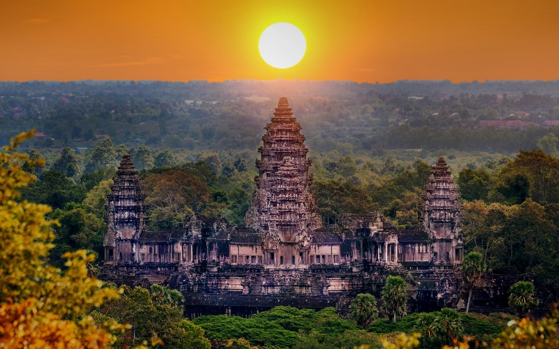 Sunset over Angkor Wat temple, with the golden sun behind and green forest surrounding.