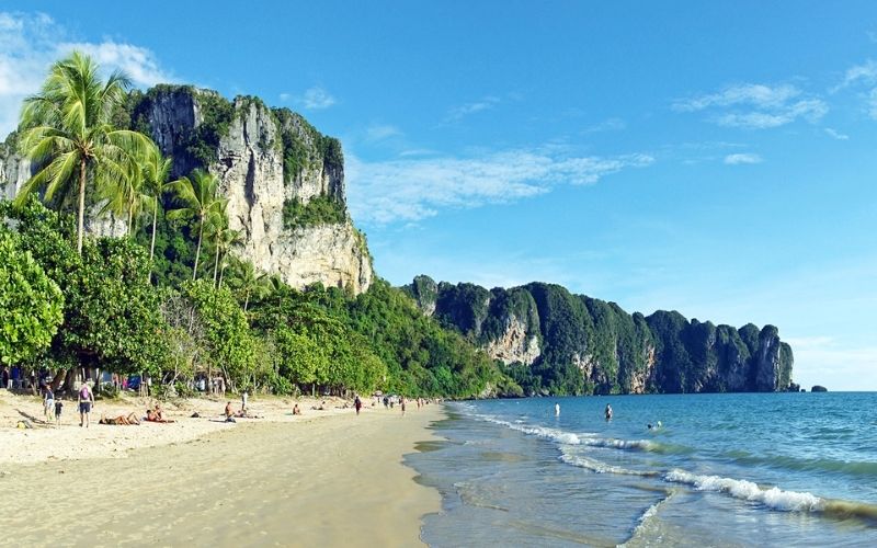 A scenic view of Ao Nang Beach in Krabi, Thailand, featuring golden sand, turquoise water, palm trees, and towering limestone cliffs under a bright blue sky.