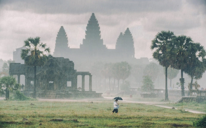 A person holding an umbrella walks through the rain in front of Angkor Wat in Cambodia, with the ancient temple and palm trees shrouded in mist.
