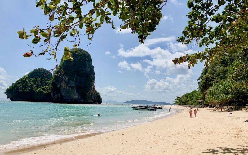 A picturesque tropical beach with soft white sand, clear turquoise water, and a prominent towering limestone karst island in the distance. Lush green foliage frames the top of the image, with people walking on the beach and boats docked near the shore under a sunny sky.