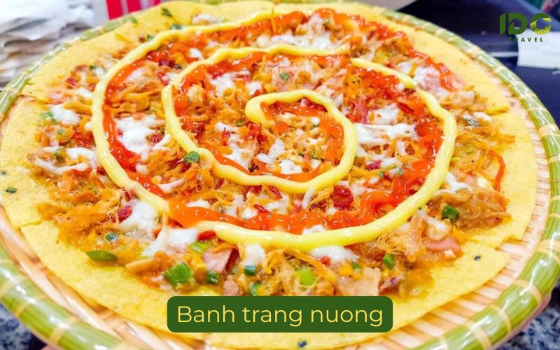 A close-up photograph of a circular Banh Trang Nuong, often called 'Vietnamese Pizza'. The thin, grilled rice paper base is completely covered with a mix of chopped green onions, dried shrimp, egg, and mayonnaise and chili sauce drizzled in a spiral pattern. It sits in a bamboo serving tray with a woven edge, with a label reading "Banh trang nuong" at the bottom. The IDC Travel logo is in the top right corner.