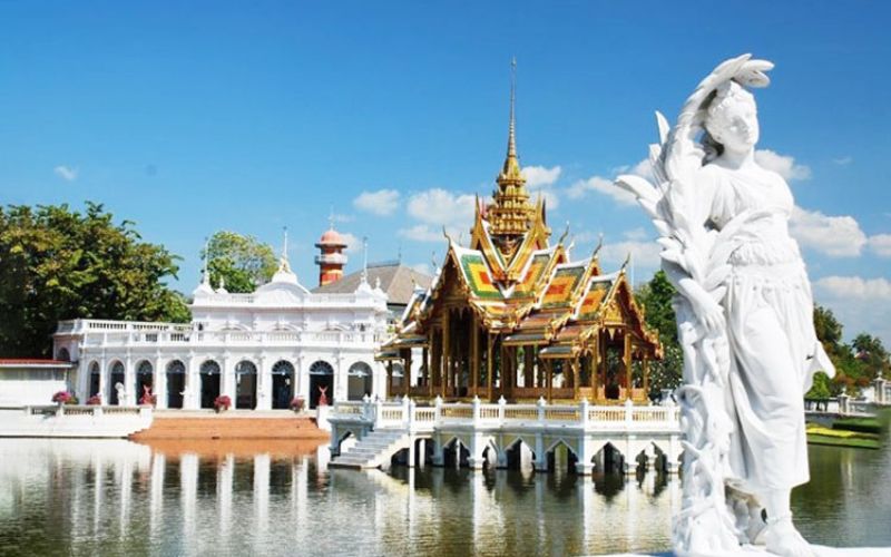 Ornate pavilion at Bang Pa-In Summer Palace in Thailand, reflected in a serene lake with a white classical statue in the foreground.
