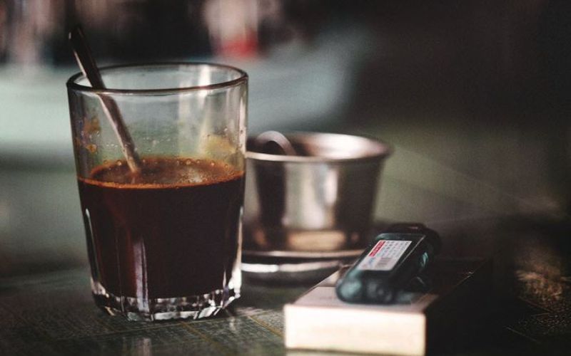 A glass of traditional Vietnamese black coffee served with ice and a metal cup, placed on a rustic table in Hanoi