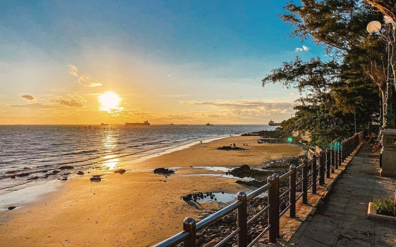A serene view of Bai Dau in Vung Tau, with calm turquoise waters meeting a rocky shoreline, lush green hills in the background, and a peaceful coastal atmosphere away from the city crowds.