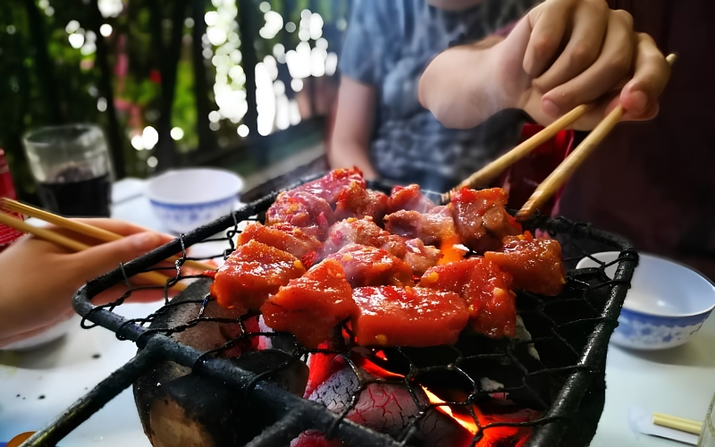 People grilling marinated beef cubes over charcoal at a table using chopsticks in Nha Trang, Vietnam.