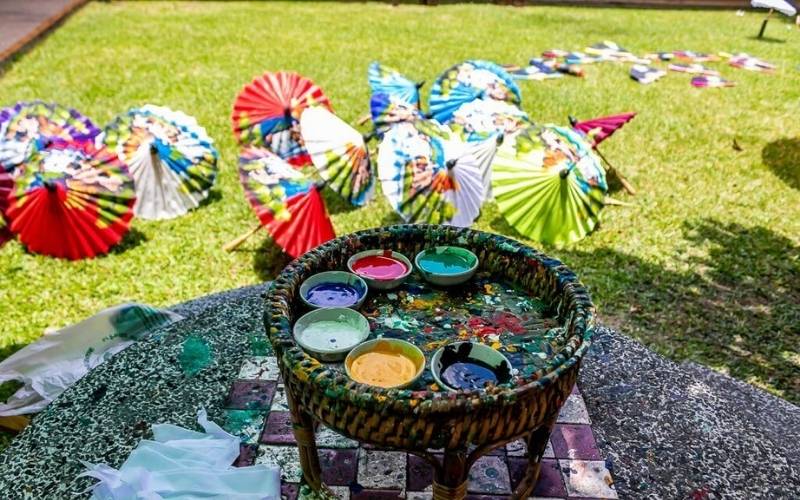 Colorful handcrafted paper umbrellas laid out on the grass to dry, with bowls of vibrant paint in the foreground at a traditional handicraft village in Thailand.