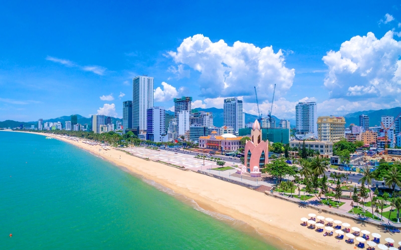 Aerial view of Nha Trang beach with clear blue water, sandy beach, and high-rise buildings lining the shore.