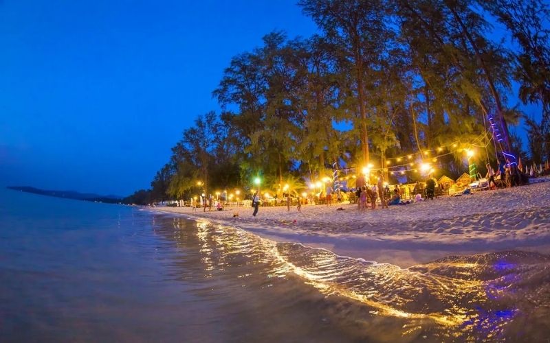 A low-angle photograph of Bang Tao Beach at twilight, featuring tall trees and sandy shores illuminated by warm yellow string lights, with gentle waves washing the sand in the foreground where people are gathered.