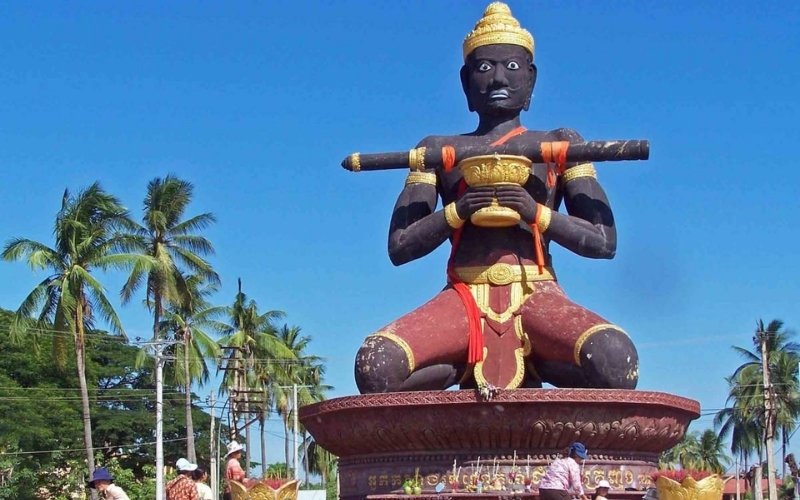 A large statue of a black guardian figure holding a club and a bowl, surrounded by palm trees under a clear blue sky in Battambang, Cambodia.