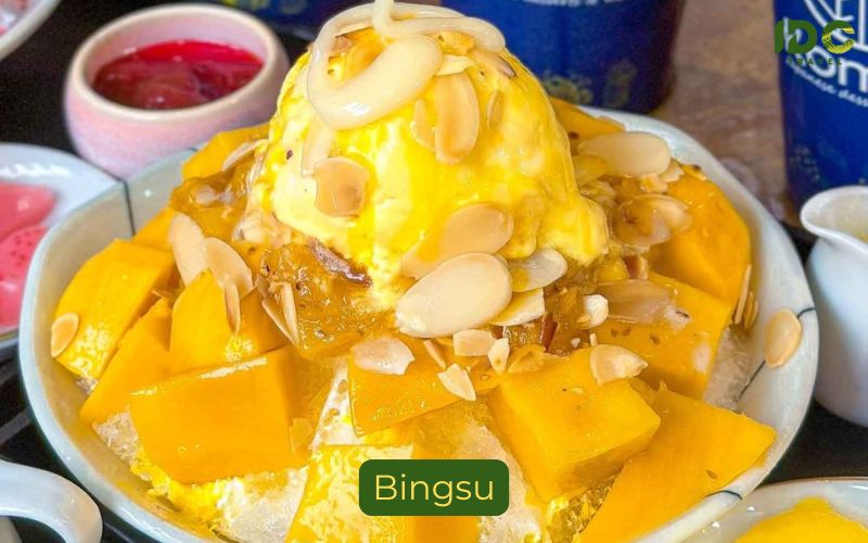 A bowl of Mango Bingsu, a Korean-style shaved ice dessert. It features a mountain of milk-flavored shaved ice topped with generous cubes of fresh, ripe mango, a scoop of vanilla ice cream, and sliced almonds. A sauce, likely mango or caramel, is drizzled over the top. Small containers of red bean paste and more almonds are in the background. A label below reads "Bingsu" with the IDC Travel logo visible.