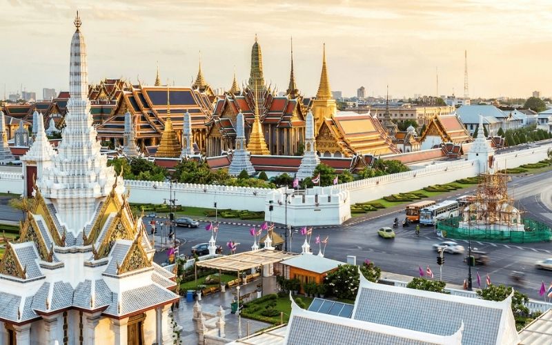 A scenic view of the Grand Palace complex in Bangkok during sunset, with golden rooftops and traditional Thai architecture glowing under a soft sky