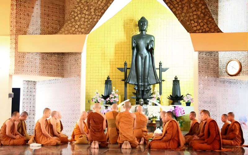 Buddhist monks sitting in a circle on the floor of a temple, praying or meditating in front of a tall, dark standing Buddha statue with a gold backdrop.