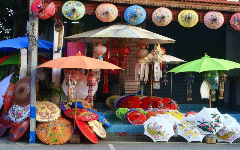A colorful market stall, likely in Bo Sang near Chiang Mai, Thailand, showcasing dozens of handmade paper umbrellas (parasols) and lanterns. Umbrellas in various sizes and vibrant colors (red, blue, yellow, orange, green) decorated with floral and bird motifs are displayed on the floor, propped up, and hanging from the eaves of the shop. Various paper lanterns are also hanging in the center.