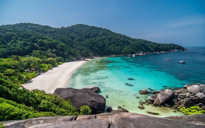 A secluded tropical beach with turquoise water, white sand, rocky shoreline, and lush green hills under a clear blue sky.