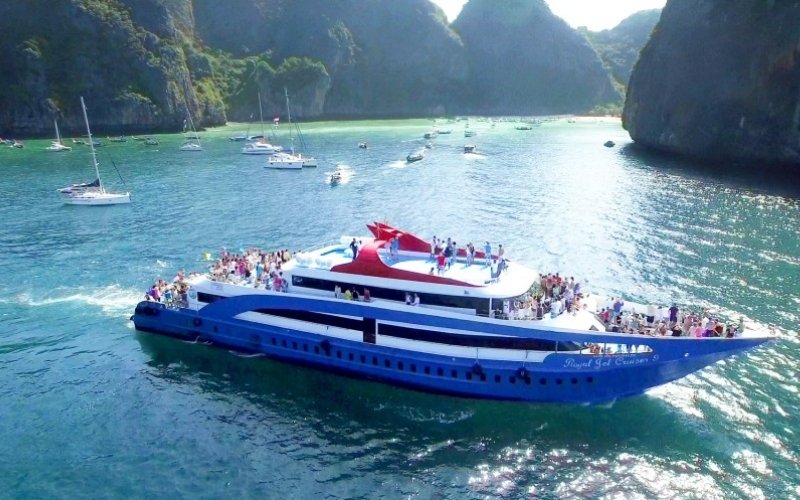 A large blue and white tour boat filled with people cruising through the turquoise waters of Phi Phi Island, surrounded by limestone cliffs and smaller boats.