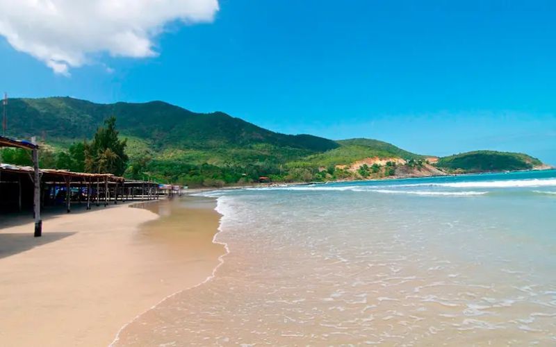Bai Dai Beach with soft sandy shoreline, gentle waves, beach huts, and green hills in the background under a clear blue sky.