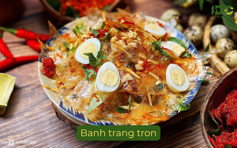A vibrant bowl of Banh Trang Tron, a Vietnamese shredded rice paper salad, presented on a wooden table. The salad is a mix of torn rice paper, sliced green mango, peanuts, dried beef, and Vietnamese coriander, topped with several halved hard-boiled quail eggs. The bowl is set among fresh red chilies, quail egg shells, a lime wedge, and a bowl of chili flakes. A label below reads "Banh trang tron" with the IDC Travel logo on top.