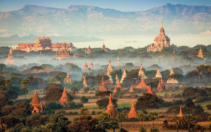 Ancient temples of Bagan, Myanmar, emerging through a soft morning mist with distant mountains and golden sunlight illuminating the landscape.