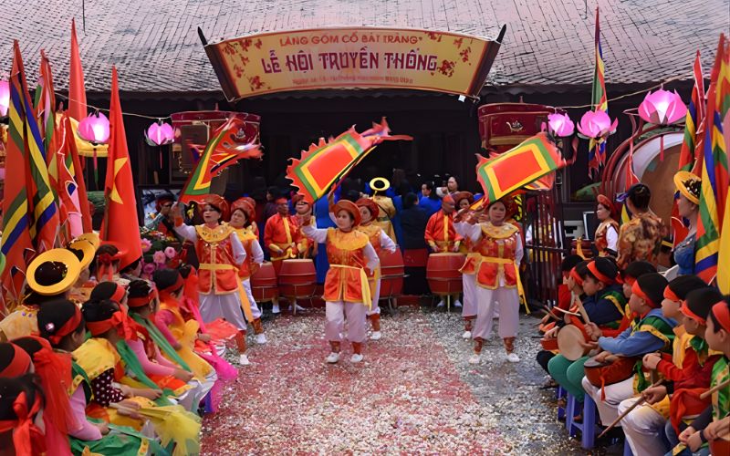 Performers in traditional orange and yellow costumes waving large festival flags during the Bat Trang Pottery Village Festival, with a decorative archway, pink lotus lanterns, and a group of children playing drums in the foreground.