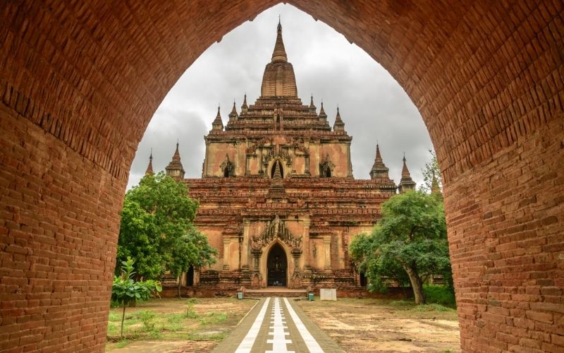An ancient temple in Bagan, Myanmar, viewed through a brick archway with symmetrical architecture, lush green trees, and a cloudy sky above.