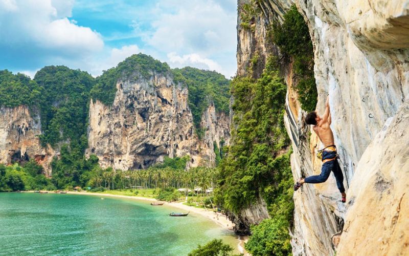 Rock climber scaling limestone cliff at Railay Beach in Krabi, Thailand, with stunning coastal scenery in the background