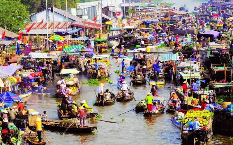 A group of people board a long wooden boat on a narrow canal in Can Tho. They are surrounded by thick green trees and low-hanging branches, with some passengers already seated and others stepping carefully from the muddy bank into the boat.
