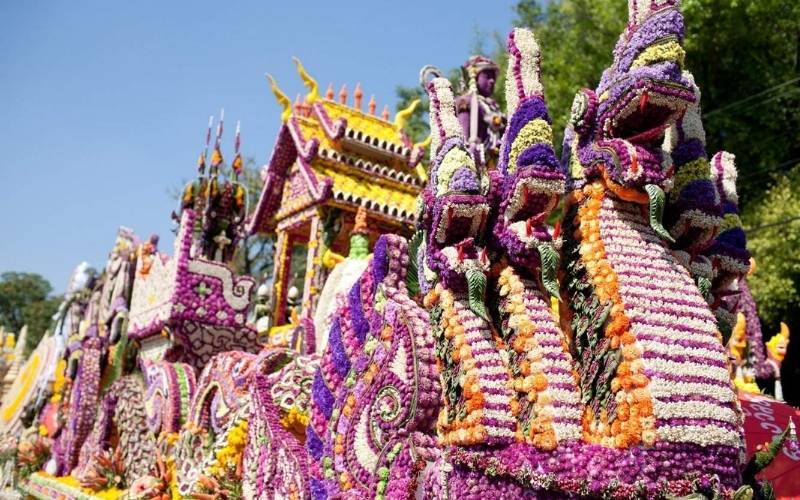 A close-up shot of an elaborate parade float at the Chiang Mai Flower Festival, Thailand. The float is intricately covered entirely in fresh flowers of various colors, predominantly purple, yellow, and white. The prominent feature is a large, serpent-like Nāga figure, sculpted from purple and white flowers, leading the float, with a traditional Lanna-style pavilion structure decorated in yellow and purple flowers visible behind it.