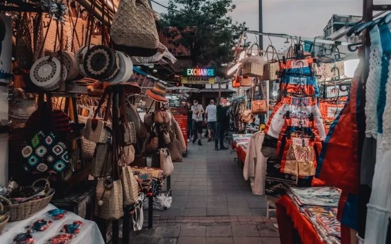 A view down a narrow, bustling aisle at the Chiang Mai Night Bazaar, flanked by market stalls overflowing with handmade crafts, including woven straw bags, hats, and patterned fabrics. An "Exchange" sign is visible in the distance, and the stalls are illuminated by warm market lights.