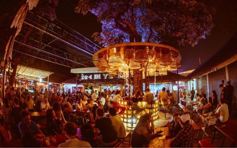 A wide-angle, lively night photograph of the bustling Zoe in Yellow/Loft Garden nightlife complex in Chiang Mai. The outdoor area is filled with people seated at tables and socializing around a circular, illuminated bar built around a large tree, decorated with woven basket lamps hanging from the canopy. Neon signs and warm string lights create a vibrant party atmosphere.