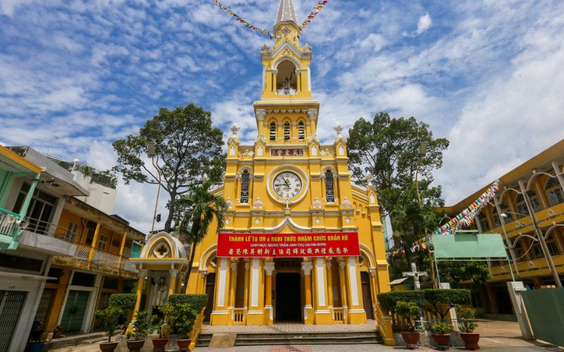 The vibrant yellow facade of Cha Tam Church in District 5, Ho Chi Minh City, featuring a tall bell tower with a clock and white trim. The building showcases a unique fusion of European Gothic design and Eastern architectural details, set against a bright blue sky with white clouds. A red banner with Vietnamese and Chinese text hangs above the entrance.