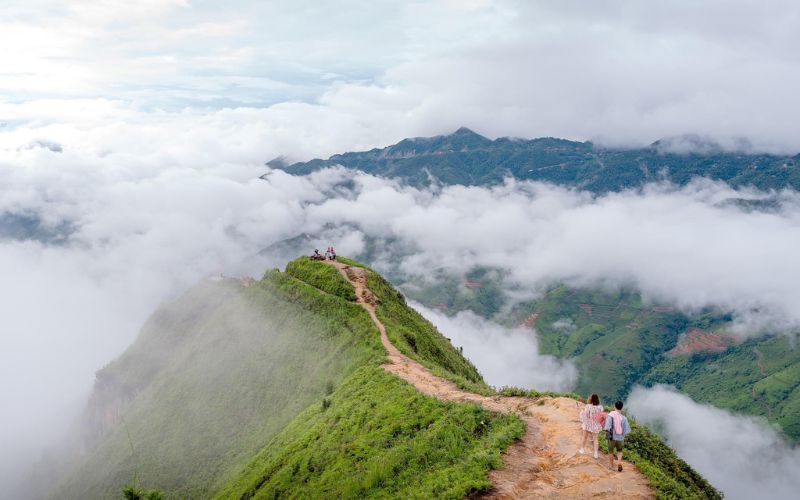 Hikers walking along the Dinosaur’s Backbone trail in Ta Xua, Vietnam, surrounded by a sea of clouds with mountain ranges in the distance