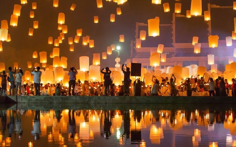 A spectacular nighttime view of the Yee Peng (Loy Krathong) Lantern Festival in Chiang Mai, showing a large crowd releasing dozens of illuminated sky lanterns over a body of water.