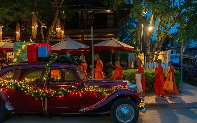Buddhist monks in orange robes walking by a vintage car decorated with Christmas lights and gift boxes during a festive evening celebration in Laos, blending traditional culture with Christmas decorations.