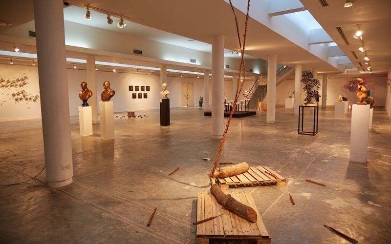 A wide shot of the spacious, minimalist exhibition hall at the Chiang Mai University Art Center, featuring various contemporary sculptures, busts on pedestals, and a raw wooden installation in the foreground.