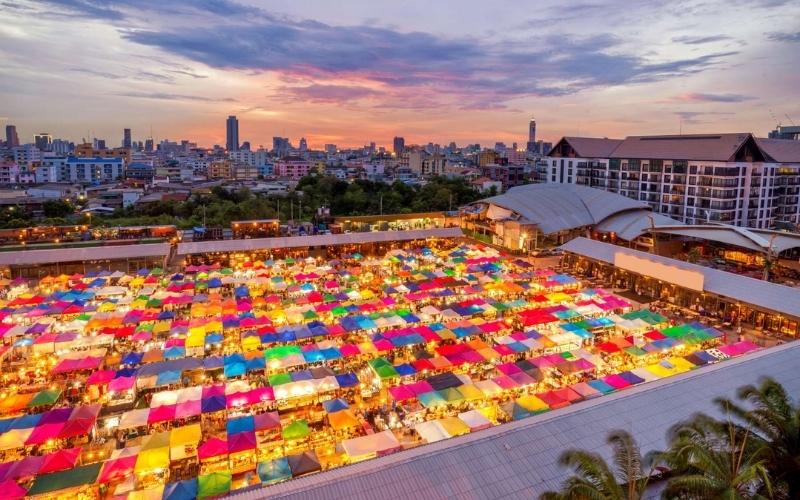 A vibrant night market with hundreds of colorful stalls seen from above, set against a city skyline at sunset.