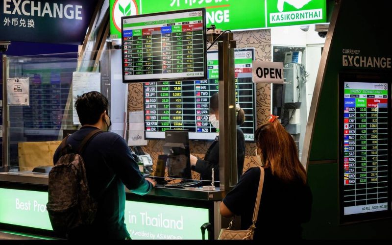 Travelers exchanging money at a currency exchange booth in Thailand, with digital rate boards displaying foreign exchange rates for various currencies