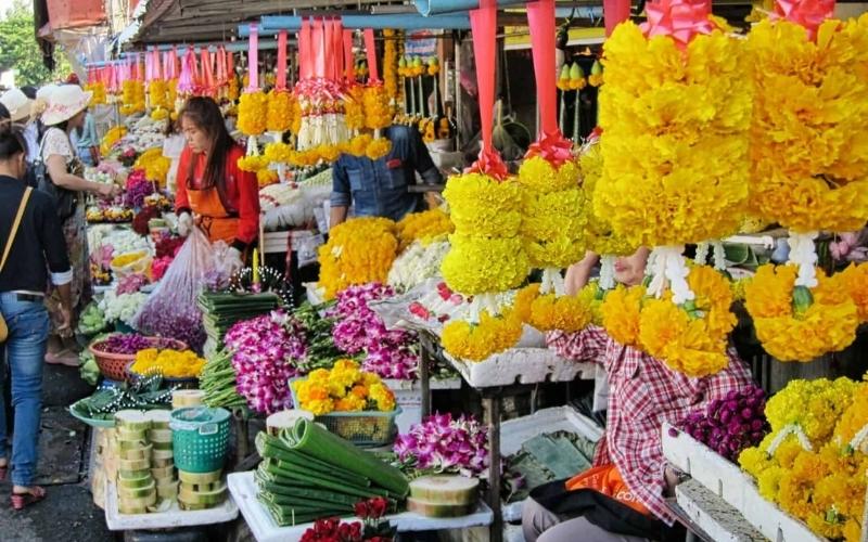 A bright photograph of the Chiang Mai Ton Lamyai Flower Market, showing market stalls overflowing with colorful hanging marigold garlands, purple orchids, and other fresh cut flowers being sold by local vendors.