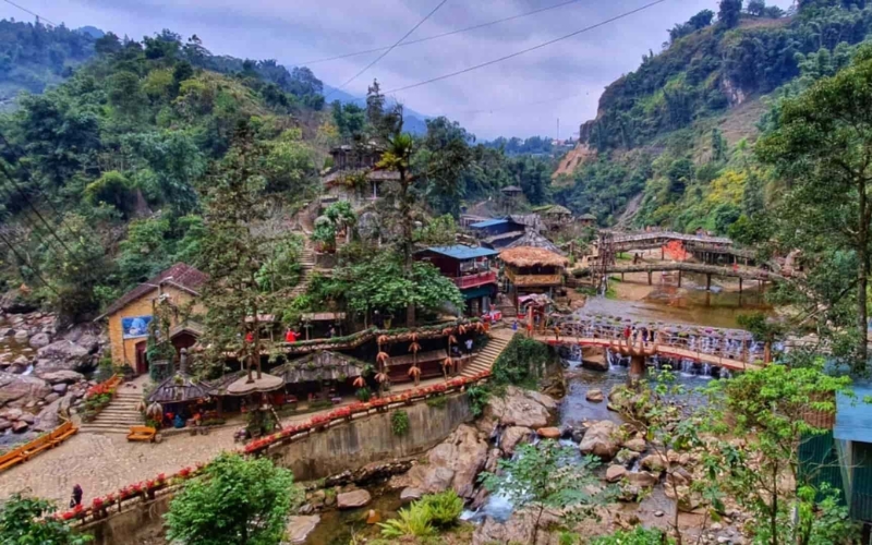Scenic view of Cat Cat Village nestled in a valley, with traditional wooden houses, bridges over a stream, and lush green hills reflecting Sapa’s cultural and natural charm.