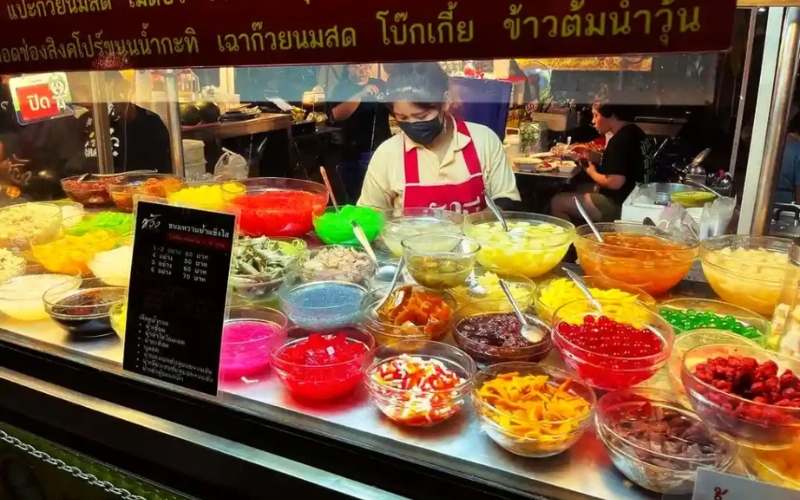 A street food vendor wearing a red apron stands behind a vibrant display of glass bowls filled with colorful jellies, fruits, and toppings for Thai desserts at Chang Phuak Gate Food Market.