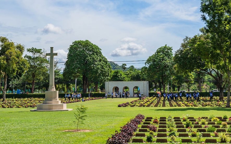 Stone cross monument standing in the center of Kanchanaburi War Cemetery, surrounded by neatly kept lawns and war graves.