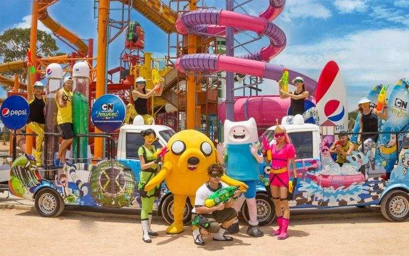 A group of staff and costumed characters, including Finn and Jake from Adventure Time, posing on a colorful float in front of the massive water slides at the Cartoon Network Waterpark near Pattaya, Thailand.