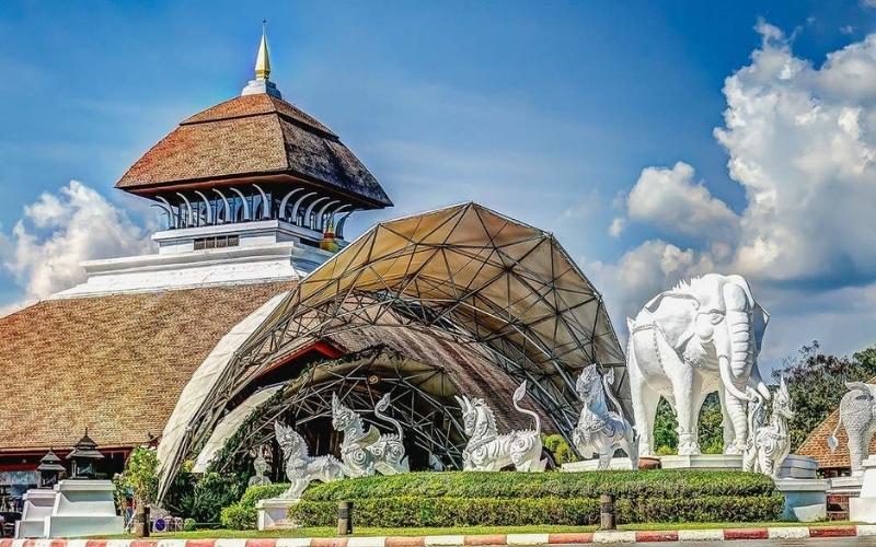 An entrance to Chiang Mai Night Safari featuring a large pavilion with a tiered roof, a geometric dome structure, and white animal statues including an elephant and mythical creatures under a bright blue sky.