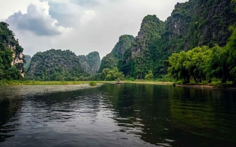 A tranquil river surrounded by lush green limestone mountains and trees under a cloudy sky, viewed from the water surface.