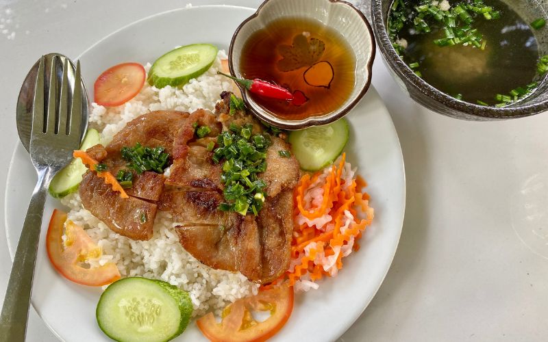 Vietnamese broken rice with grilled pork chop, pickled vegetables, cucumber, tomato, and fish sauce served with clear soup.