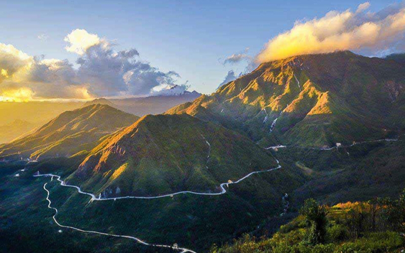 The winding mountain road of O Quy Ho Pass bathes in golden sunset light, with impressive peaks and cloud-covered ranges creating a peaceful and scenic view in Sapa.