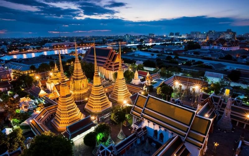 An illuminated Thai temple complex at dusk, with golden spires, ornate rooftops, and surrounding city lights reflecting off a nearby river under a deep blue evening sky.